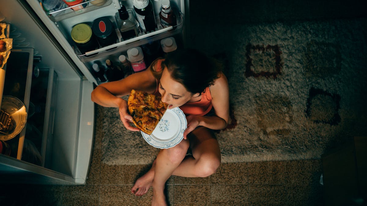 Une femme mange une pizza froide devant son frigo