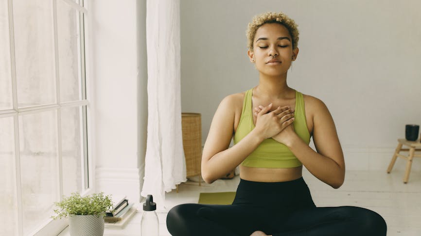 Jeune femme afro pratiquant le yoga en leggings assise sur le sol en posture de lotus mains croisées sur la poitrine (au niveau du nerf vague) léger délice sourire sur le visage, sentant son corps. 