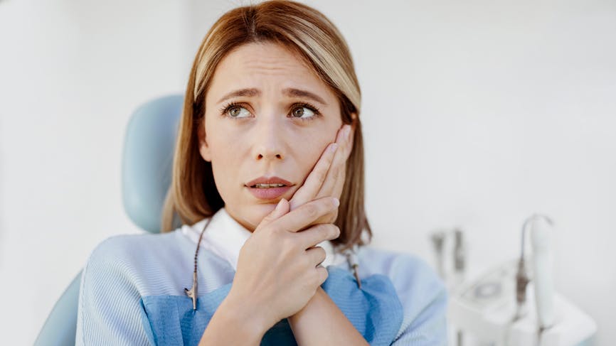Femme qui semblent se plaindre de maux de dents dans un cabinet dentaire. Elle est assise sur le siège du dentiste et se tient la joue. 