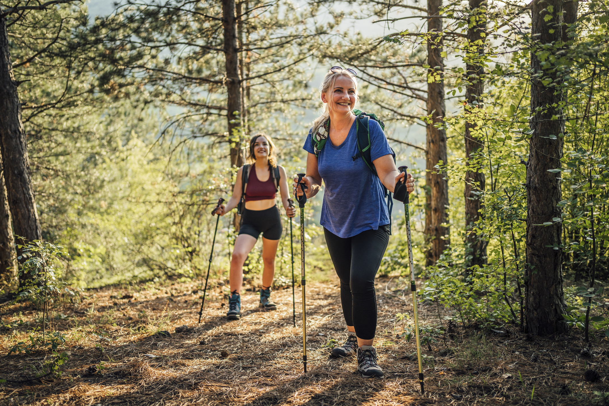 Deux femmes font de la randonnée avec des bâtons dans une forêt ensoleillée.
