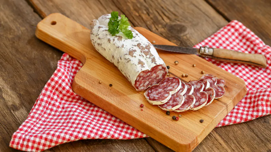 Saucisson tranché sur une planche en bois avec couteau et nappe à carreaux rouges.