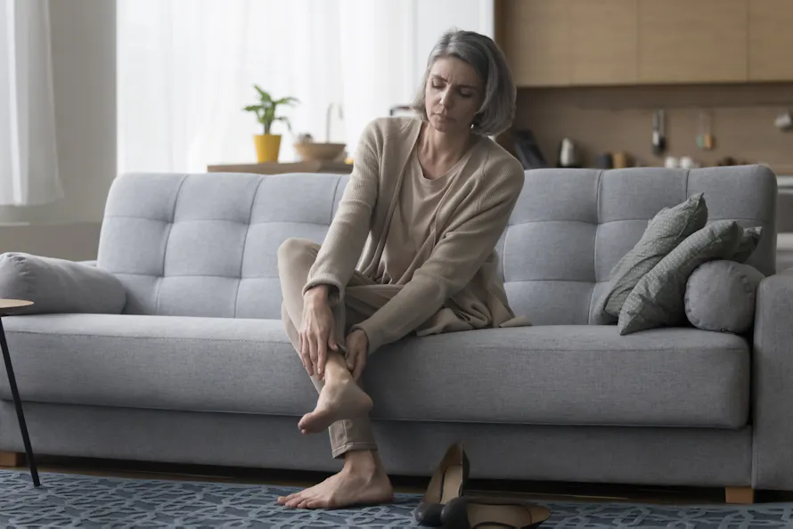 Une femme assise sur un canapé, massant sa cheville dans un salon lumineux.